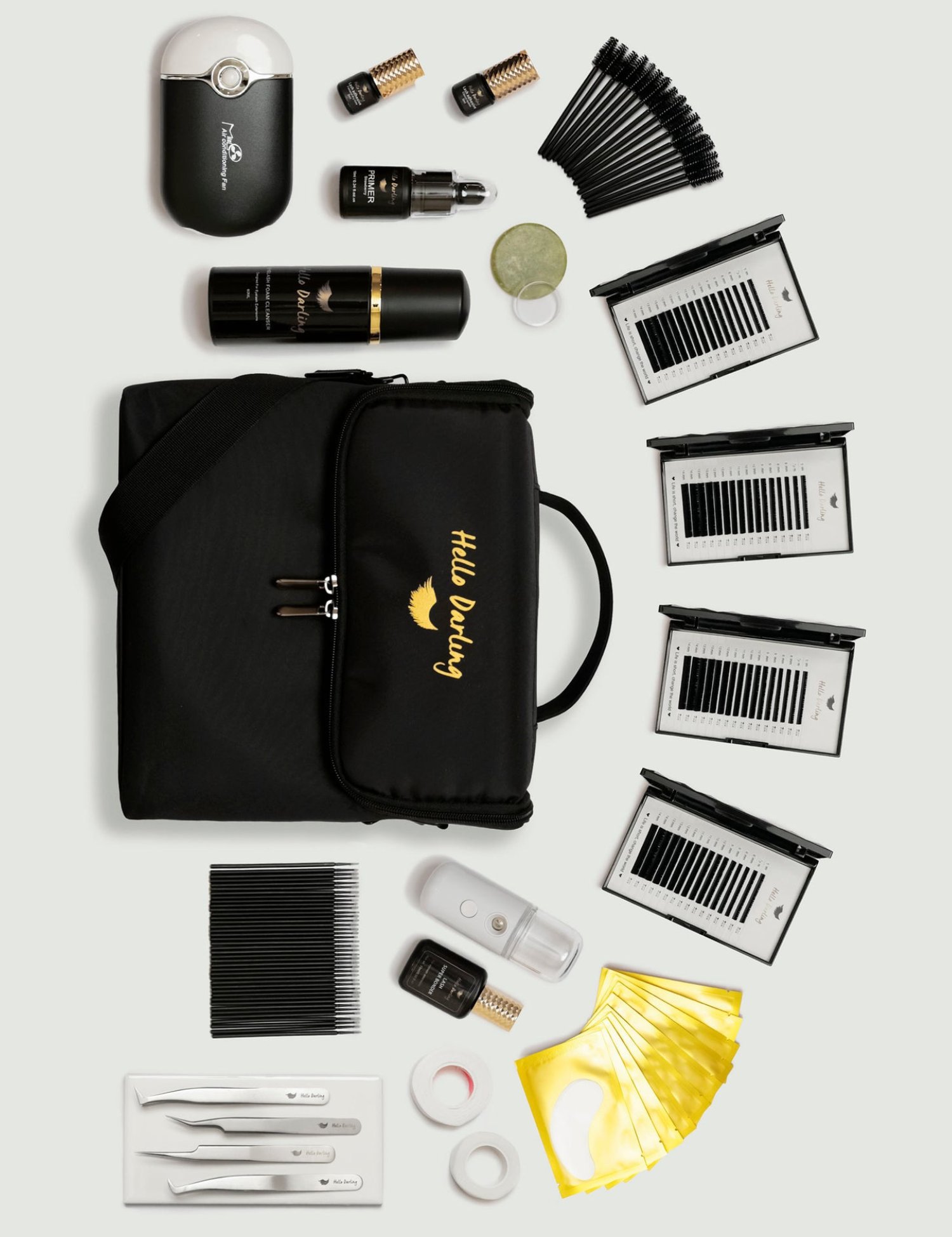 A meticulously organized assortment of eyelash extension supplies laid out on a white surface. The collection includes a black motorized fan, multiple boxes of varying lash extensions, black and gold branded lash primers and cleaners, and assorted tools like tweezers and brushes, all arranged around a central black carrying case with 'Hello Darling' embroidered in gold.