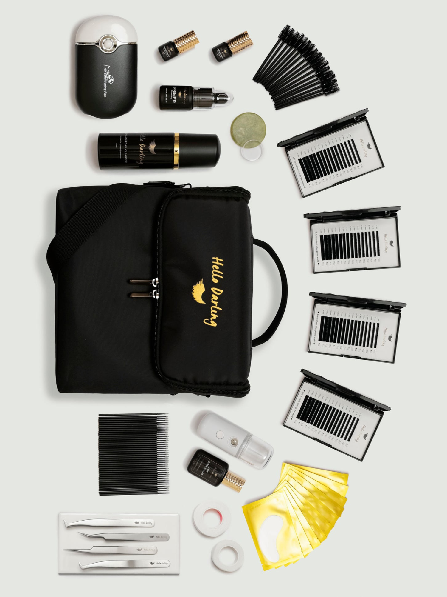 A meticulously organized assortment of eyelash extension supplies laid out on a white surface. The collection includes a black motorized fan, multiple boxes of varying lash extensions, black and gold branded lash primers and cleaners, and assorted tools like tweezers and brushes, all arranged around a central black carrying case with 'Hello Darling' embroidered in gold.