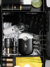 Close-up view inside a black organizer filled with eyelash extension tools and products. Items include a sleek black motorized fan, lash primer, lash cleanser in black and gold bottles, various tweezers, and lash extension trays neatly arranged in a padded compartment, emphasizing precision and professional presentation