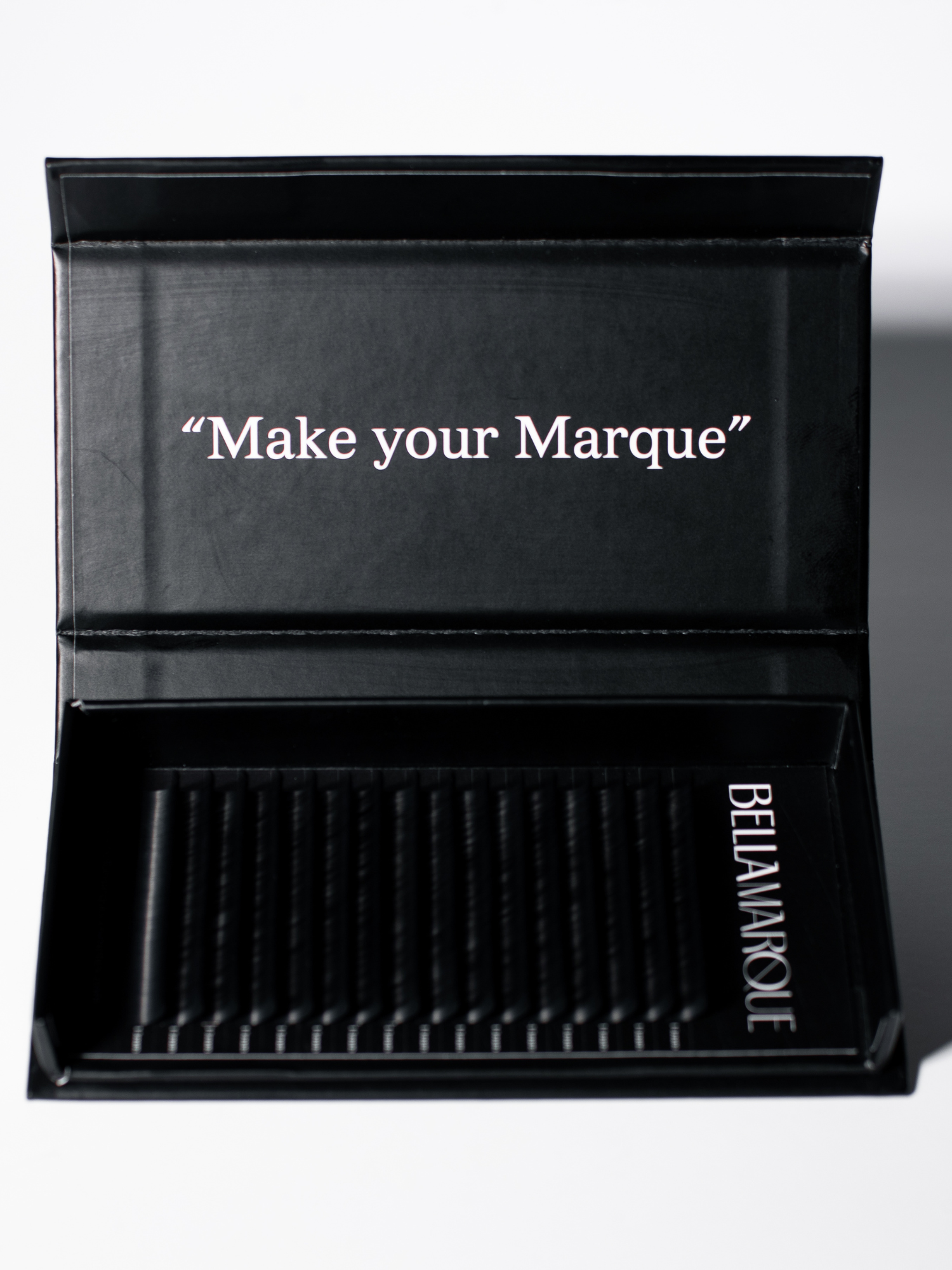 Open lash extension tray displaying long, black synthetic lashes neatly aligned, with the slogan 'Make your Marque' in white cursive script inside the lid.