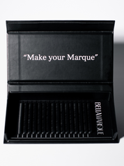 Open lash extension tray displaying long, black synthetic lashes neatly aligned, with the slogan 'Make your Marque' in white cursive script inside the lid.